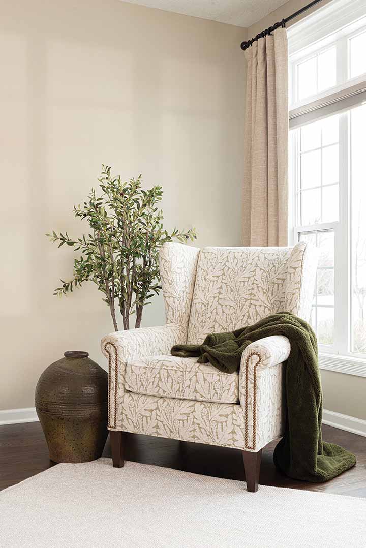 SmithBrothers_Home_Chair_144dpi A tan colored upholstered chair with a cream colored leaf pattern sits by a brightly lit window with a decorative tree and large vase.