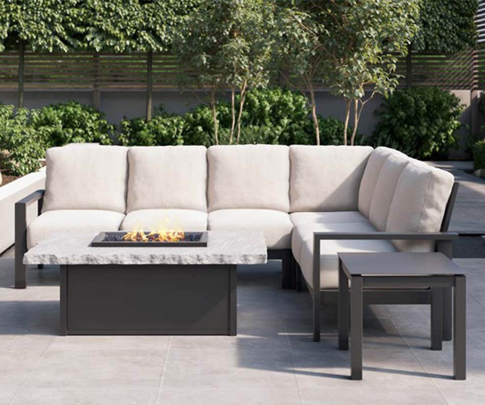 homecrest_modern_patio-furniture_uphostered-outdoor-section_marble-fire-table_brown-side-table An outdoor, dark aluminum framed sectional with grey cushions, matching side table, and table with a rectangular fire pit in the middle sits outside on a concrete patio