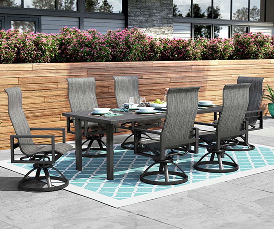 A display of outdoor patio high-backed furniture with dark metal accents and long metal table sits by a wooden wall.