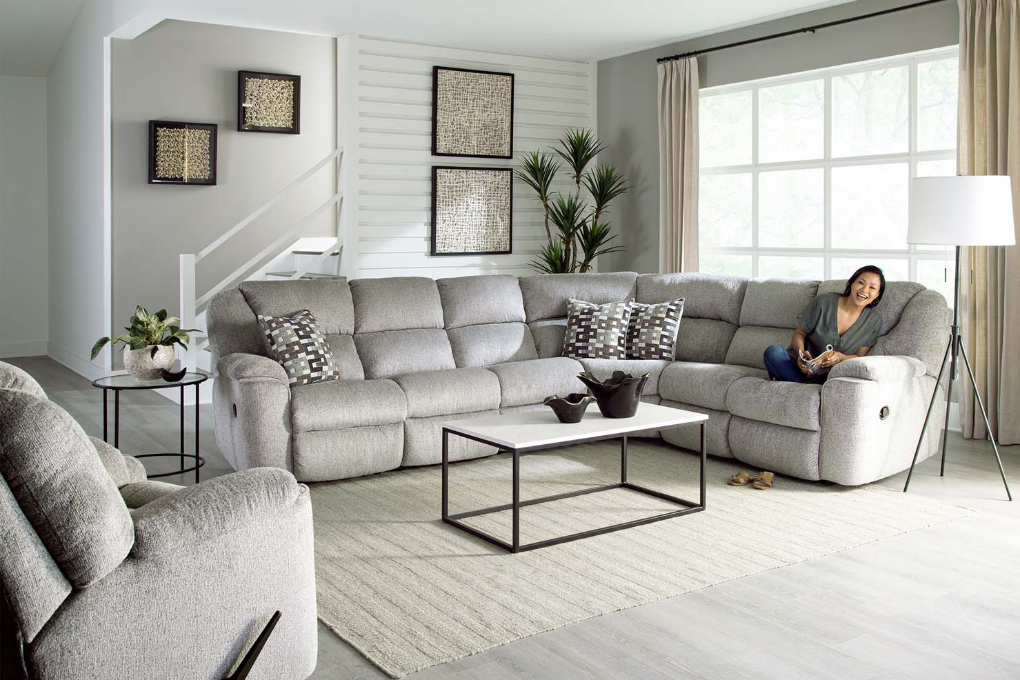 A woman smiles sitting on a light grey upholstered sectional with decorative pillows and a matching recliner facing it.