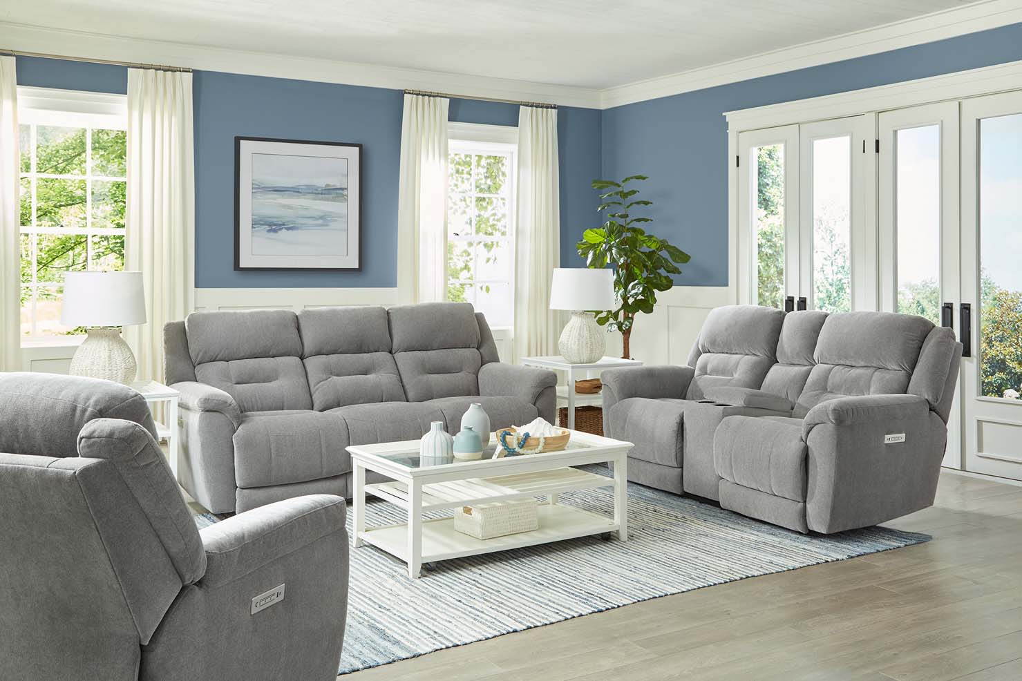 A set of light grey couch, loveseat, and chair sit inside a naturally lit living room with blue walls.