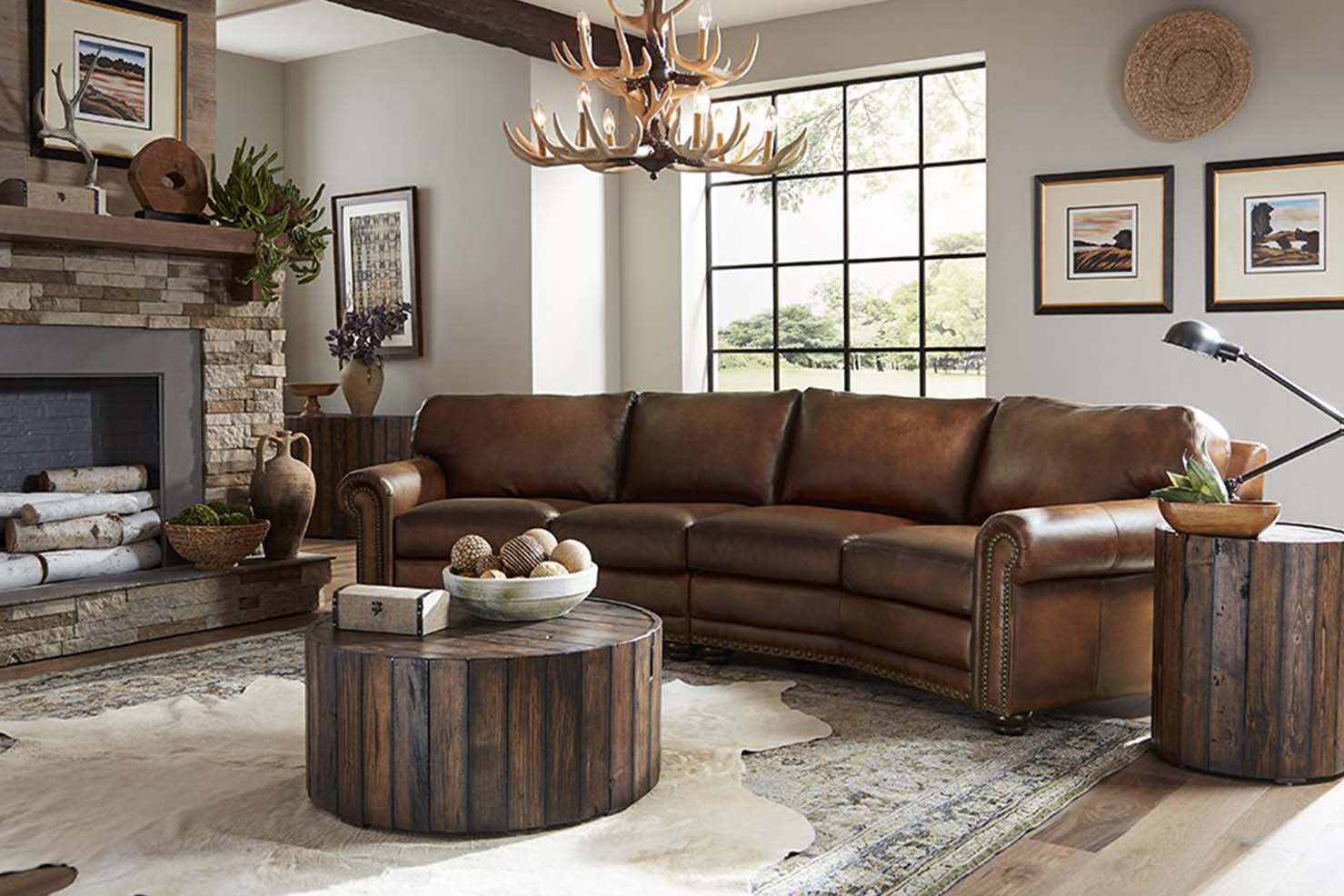 A slightly curved sofa is upholstered in a dark patina leather with bronze nail heads. It sits in a nature-inspired living room with stump-like side tables and antler chandelier.