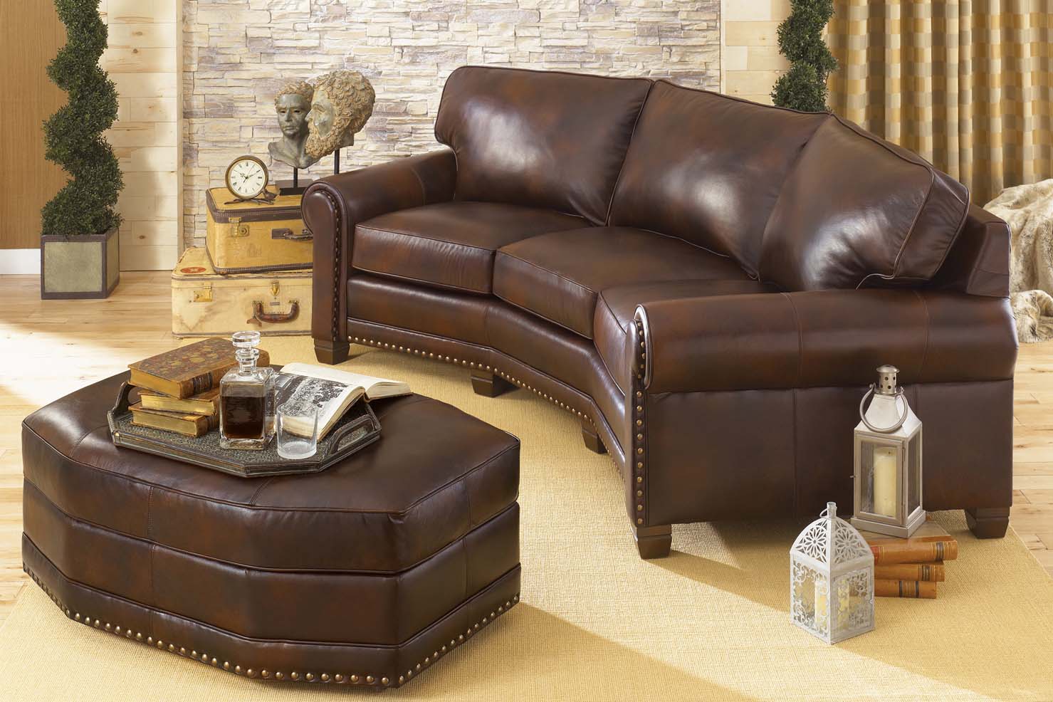 A slightly curved couch with a matching ottoman is upholstered in a shiny dark brown leather secured by golden nail heads.