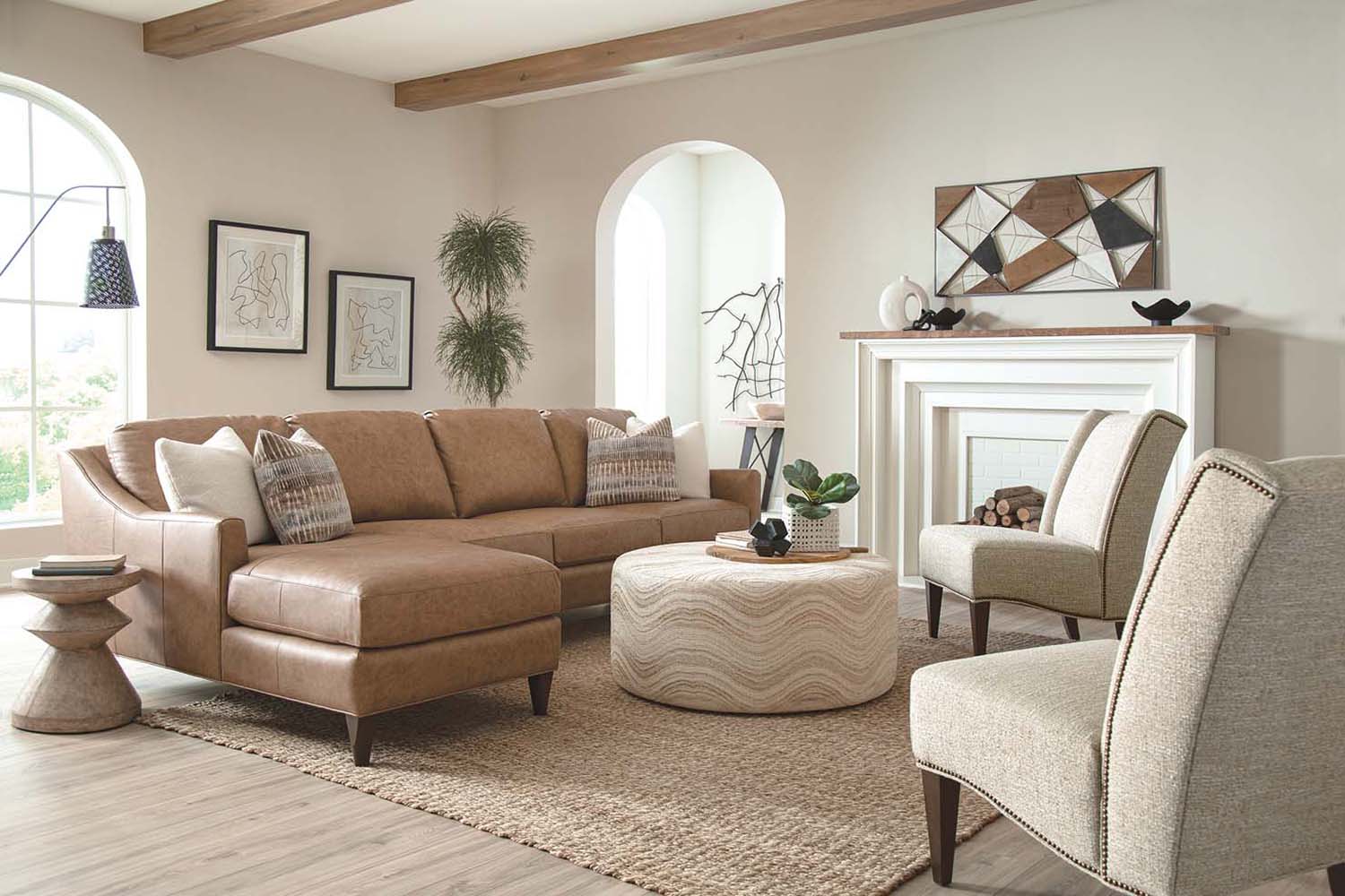 A light brown leather sectional is showcased in a beige colored living room with two fabric chairs and a patterned ottoman.