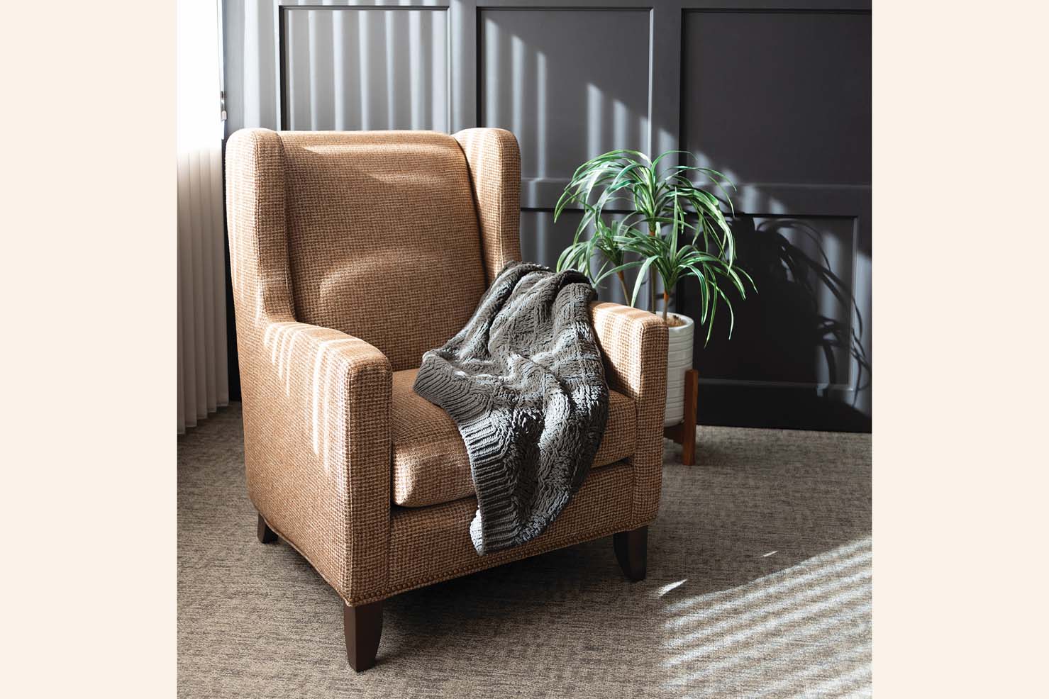 A winged occasional chair is upholstered in a textured tan fabric with a grey knitted blanket thrown over the seat.