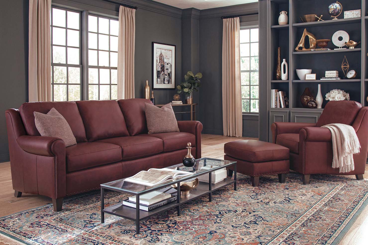 Deep red leather sofa, chair, and ottoman is displayed in a dark grey painted living room. A large patterned rug and glass coffee table accent the furniture.