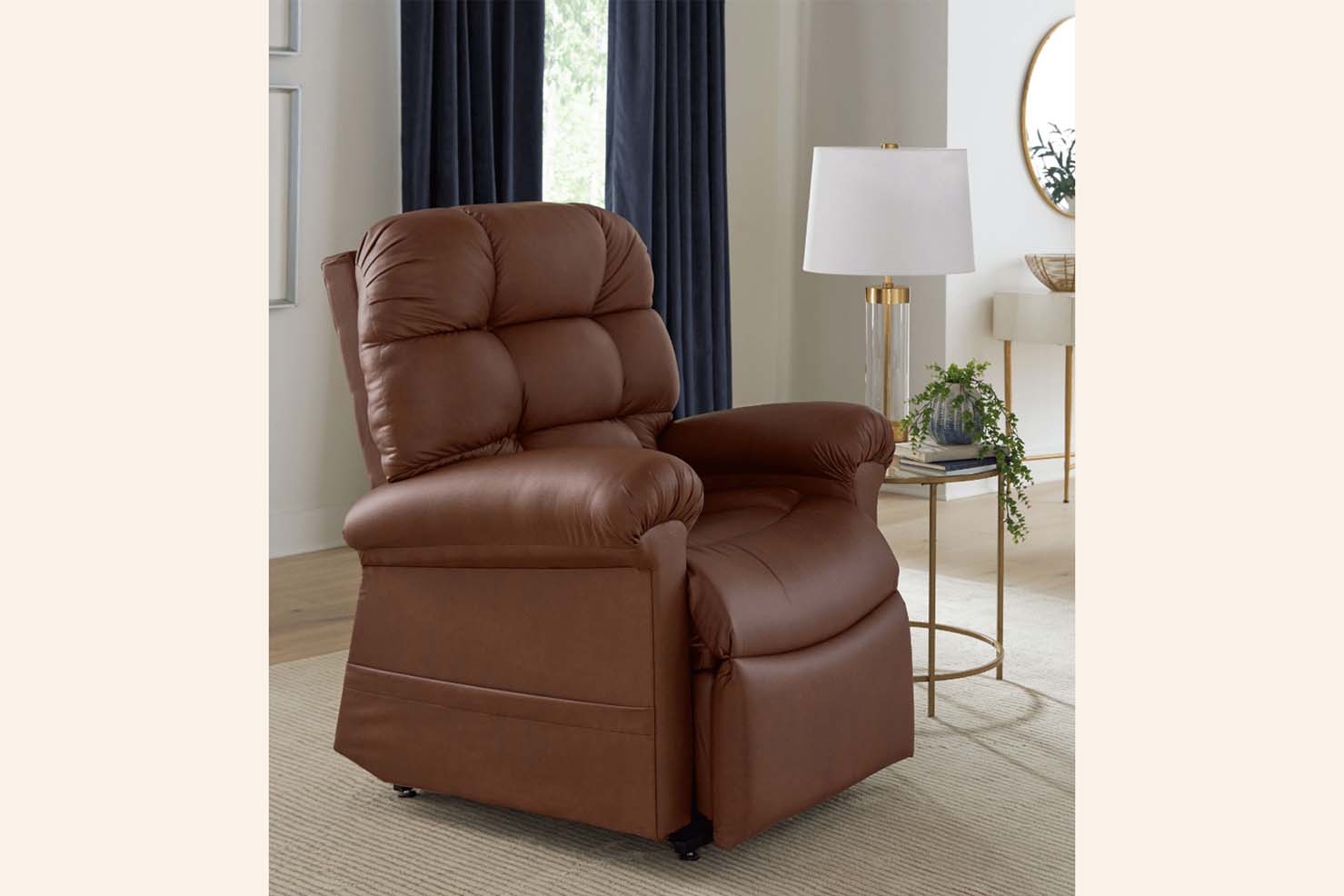 A photo of a tufted, brown leather recliner sitting in front of a window with navy drapes. A side table with a lamp and decorative plant sits besides the chair.